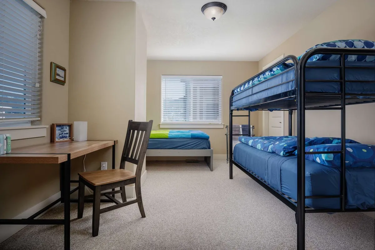 A cozy bedroom designed with bunk beds, a desk and chair, and a window with blinds. The playful yet functional setup provides ample space for children or teens to sleep, study, and play.