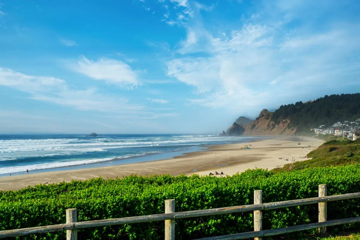 A wide view capturing the sandy shoreline, rolling ocean waves, distant cliffs, and a coastal town framed by a lush green hedge. The scene is tranquil, with clear skies and the inviting expanse of the beach ideal for family activities like beachcombing and picnicking.