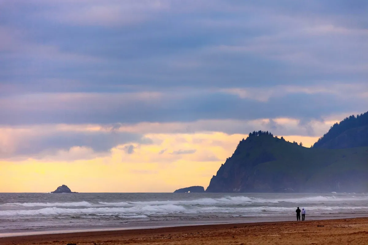 A sweeping view of the coastline with gentle ocean waves, distant cliffs, and two figures enjoying a leisurely stroll along the beach. The atmosphere is tranquil and romantic, evoking the experience of exploring the local area after a meal.