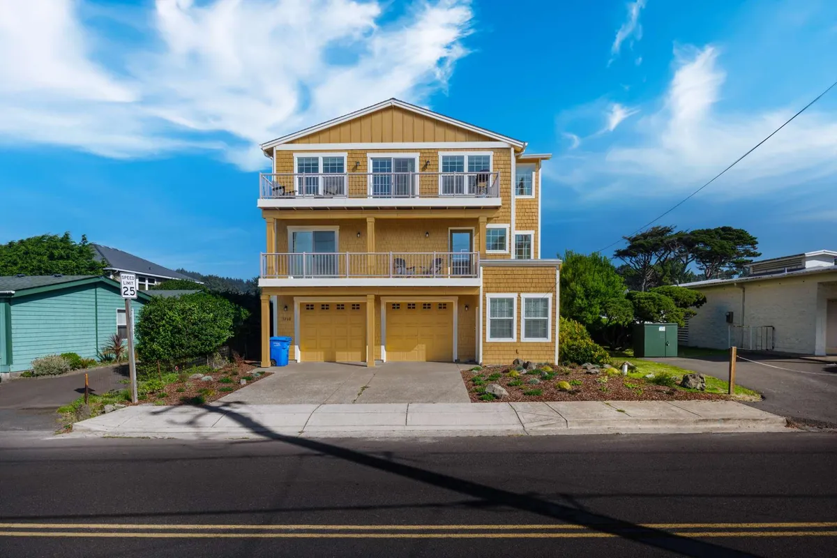 The image features a three-story coastal home with multiple balconies overlooking a beautifully landscaped garden. The house is set against the backdrop of a tranquil Lincoln City neighborhood, and the scene captures the golden glow of sunset, with families enjoying the outdoor spaces and relaxing together.