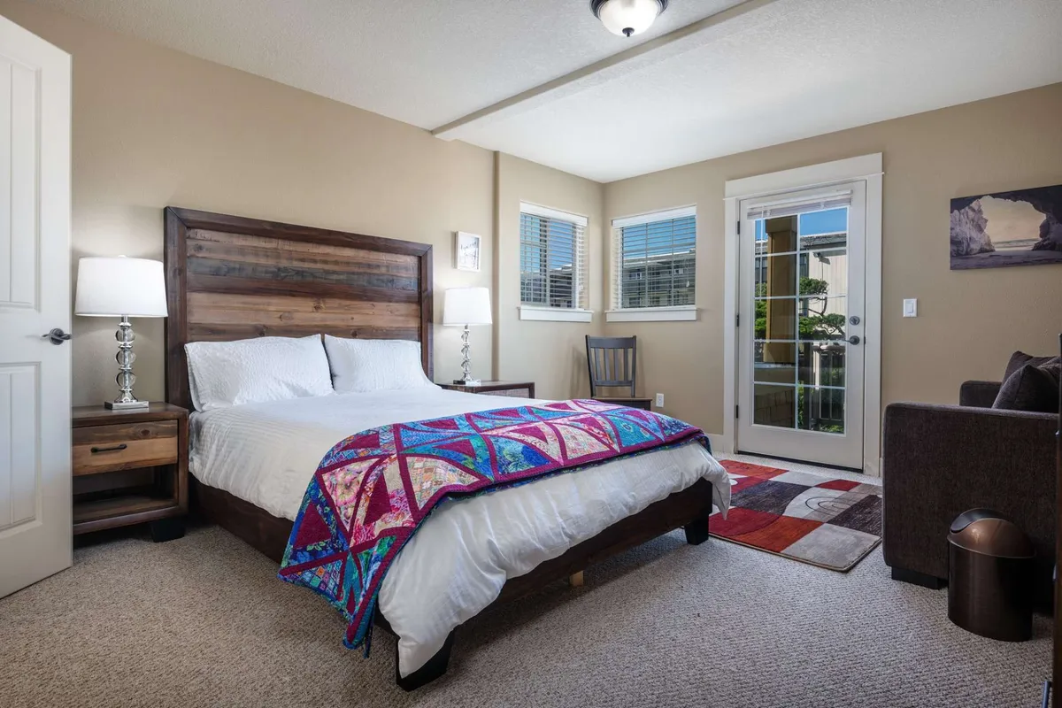 A spacious, comfortable bedroom featuring a bed with a colorful blanket, wooden headboard, matching side tables with lamps, an armchair for lounging, and doors that lead outside—inviting in natural light and offering private balcony access.