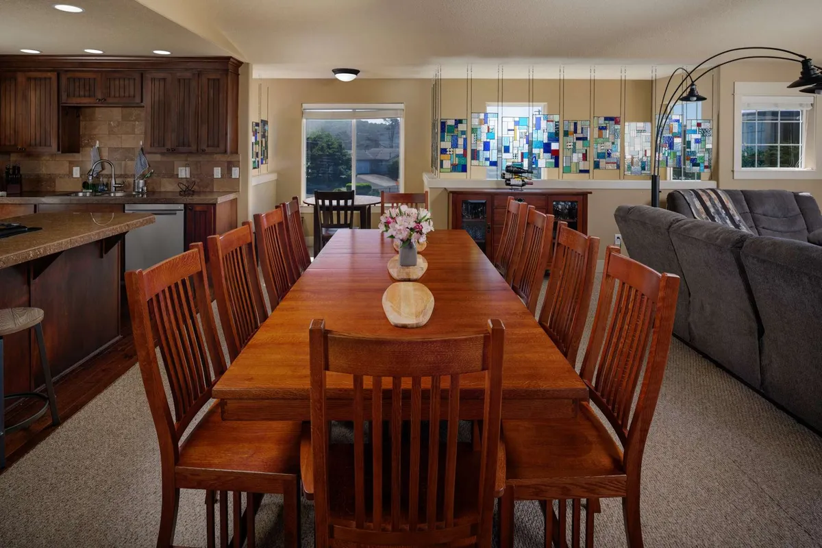 A warm, sunlit dining area featuring a long wooden table set for ten, surrounded by stained glass windows and a vibrant flower vase as the centerpiece. The space is inviting and clearly designed for group meals, celebrations, and intimate gatherings.